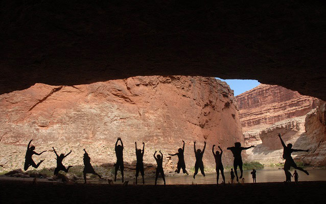 Eleven students within Redwall Cavern jumping in silhouette against cliff and river background.