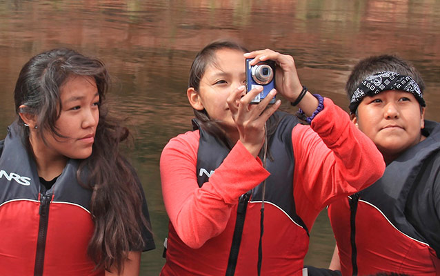 Three students standing side-by-side. The middle student is taking a photo with a small digital camera,
