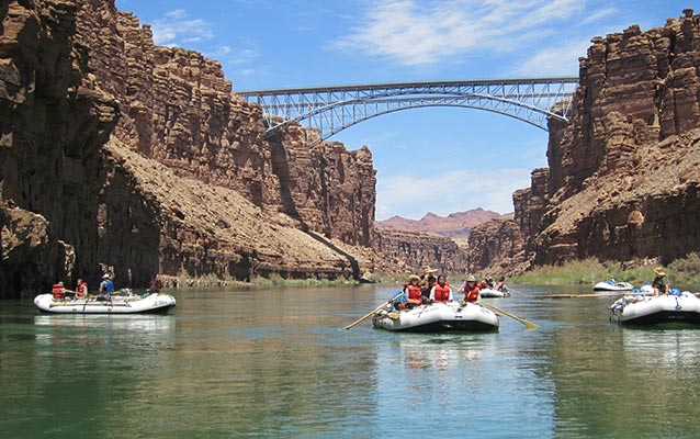 Students in four boats floating down the Colorado River with the Navajo Bridges spanning the river in the distance.
