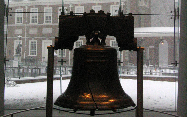 The Liberty Bell sits in the foreground with Independence Hall behind on a snowy day.