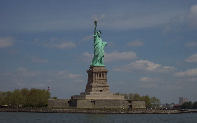 Liberty Island from a boat; the patina green statue stands on her granite pedestal on Liberty Island with blue sky behind and dark blue water below.