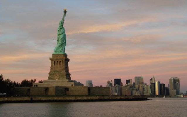 Statue of Liberty with the NYC skyline; Pink and yellow clouds reflect sunset colors.