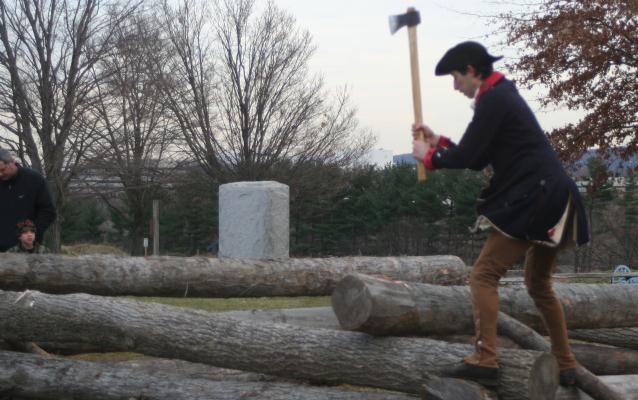 A reenactor portraying a continental soldier is swinging an axe to cut a notch in a log for a cabin