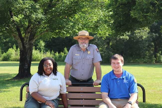 Park Ranger stands behind bench with a volunteer and intern.