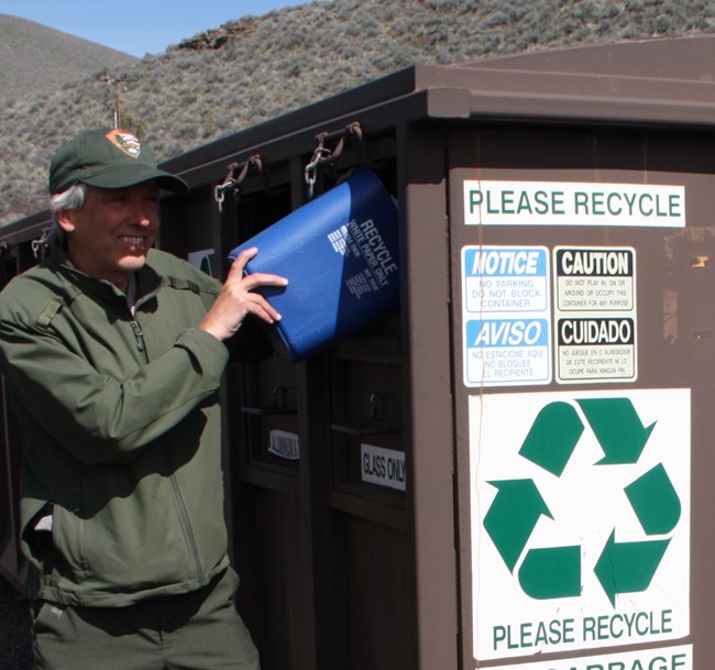 a ranger in a green uniform and hat emptying a small recycling bin into a large recycling container