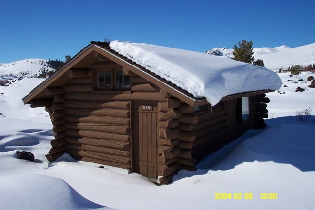 a small, brown log building completely surrounded by snow in the winter with one visible door baring a sign with the text "men"