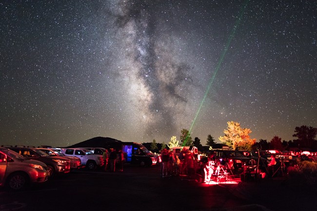 a star party illuminated by red lights while someone points at the sky with a green laser. The milky way is visible and the sky is filled with thousands of stars.