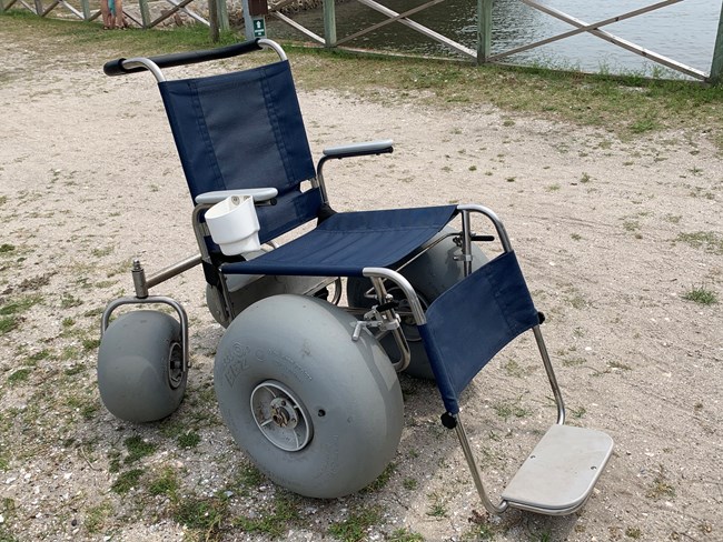 A stainless steel wheelchair with blue fabric seat and balloon tires on a sandy road