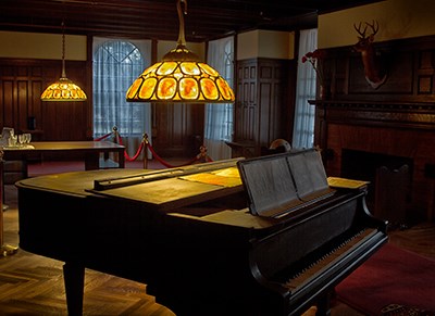 Room with dark oak wall coverings and coffered ceiling; piano under glass shaded chandeliers; deer head on wall