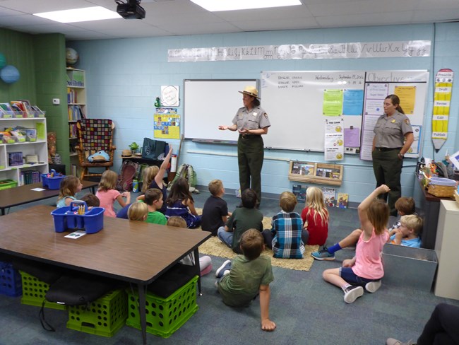 Two park rangers stand in a classroom presenting a program to children seated on the floor.