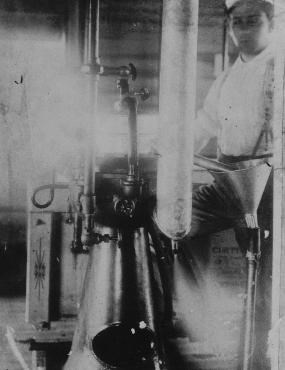 Black and white photo of a worker in Sumner Creamery in 1898. A young man in a white uniform stands in front of tubes and filters, concentrating on his work.