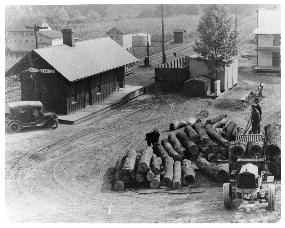 Black and white photo of the Brecksville Depot, 1932. A station building with an A-frame roof and a model-T car sit next to the tracks. A large pile of logs is in the foreground with three workers near the pile. A house and fence is in the background.