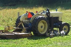 A simple tractor on a green field with cutting equipment hooked to it. Four or five chickens are gathered around it .