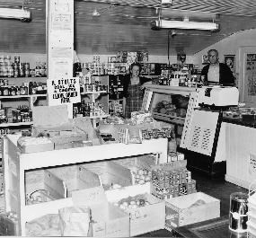 Black and white photo of Carter's General Store, Everett. Canned goods line shelves along the wall, two people stand behind a refridgerated case with a windowed front, and stacked crates create a middle island of merchandise.