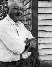 Black and white photo of Jim Szalay in 1931. A late-middle aged white man in a white button-up shirt tucked into dark pants. He is smiling and has his arms crossed over his stomach. He wears glasses and a trimmed mustache.