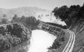 Historic photo of stream train going down railroad tracks alongside a tree lines slow moving river.