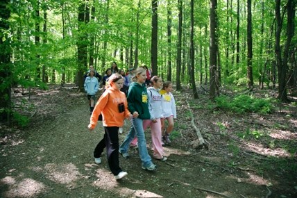 Four girls wearing colorful sweatshirts walk through a forest at the front of a larger group of students.