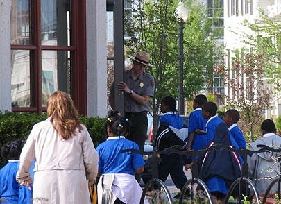 A ranger with a tan flat hat stands in a door entrance and speaks to children in blue shirts as they walk up to enter the building.