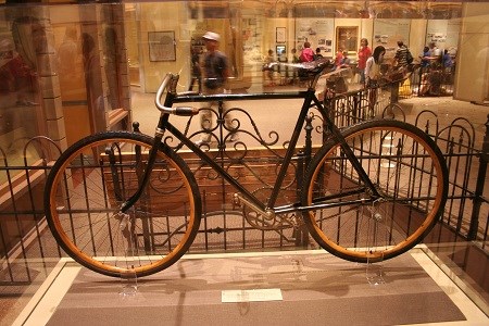 An early period bicycle on display inside of a glass case, surrounded by iron gates as people walk by.