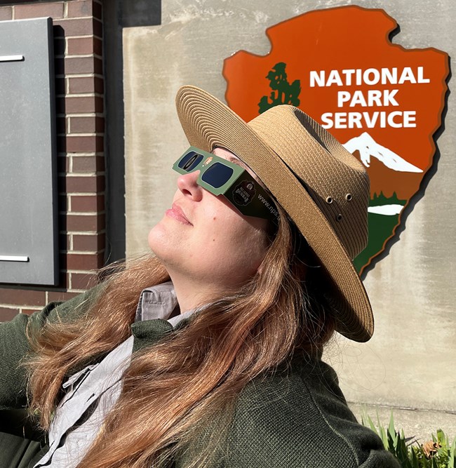 A NPS ranger looks skyward through eclipse glasses. A park sign and arrowhead are in the background.