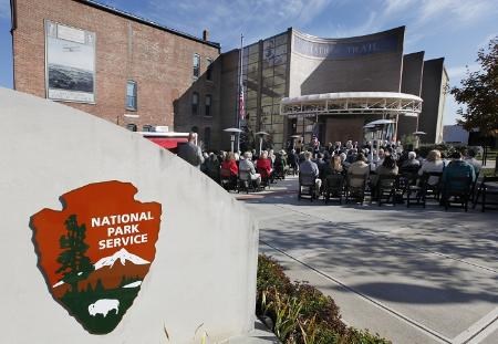 Several people seated in an open-air plaza in front of a large brick building with a sign of the NPS arrowhead logo in the foreground.