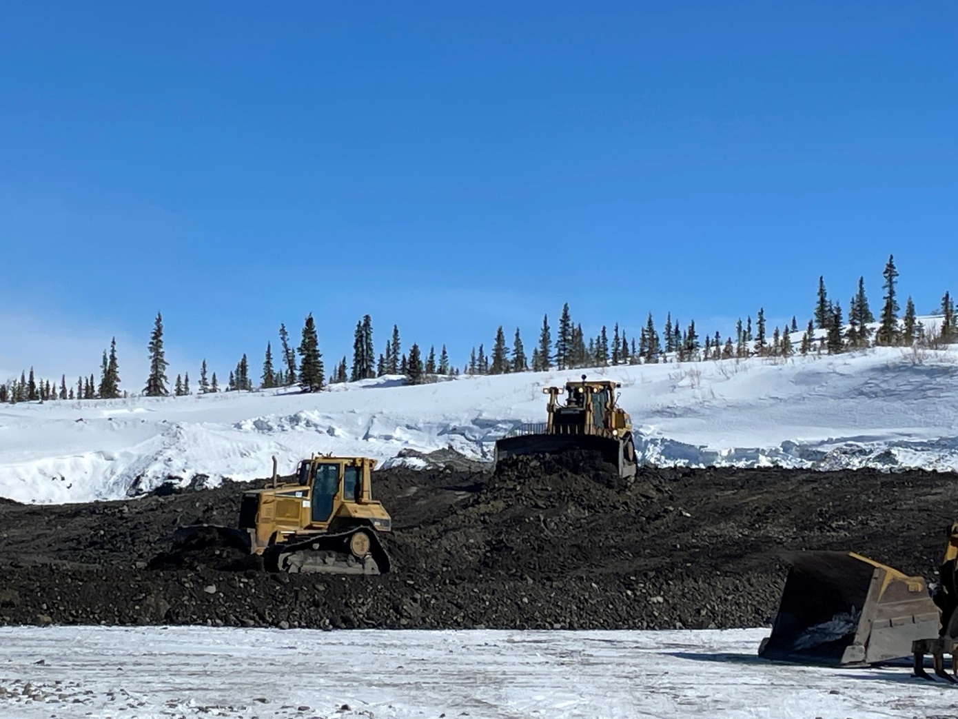 Three pieces of heavy machinery move gravel and snow to grade a large open pit.