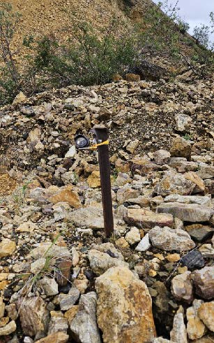 A metal post, buried in rocky ground, with a small device mounted on top of the post.