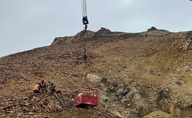 A metal box that appears to be suspended by a crane from above rests on the edge of a rocky slope. Three construction workers work on the rocks to the left.