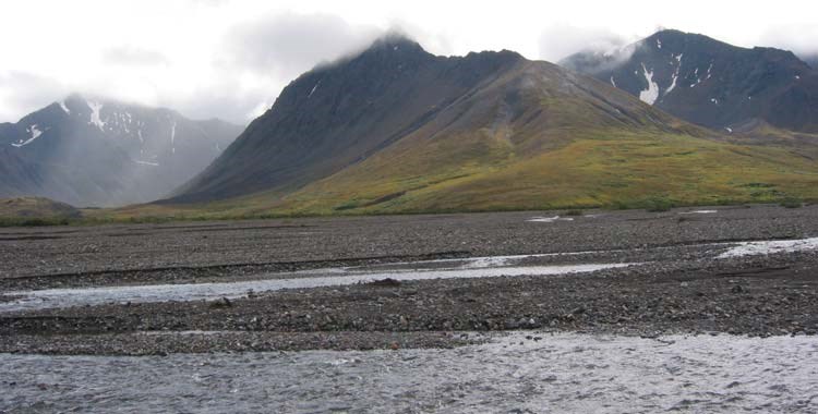a river running through a rocky plain in front of cloud-shrouded mountains