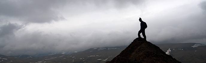 Silhouetted hiker on a rocky outcrop, misty skies and snow-dotted plain in the distance below.