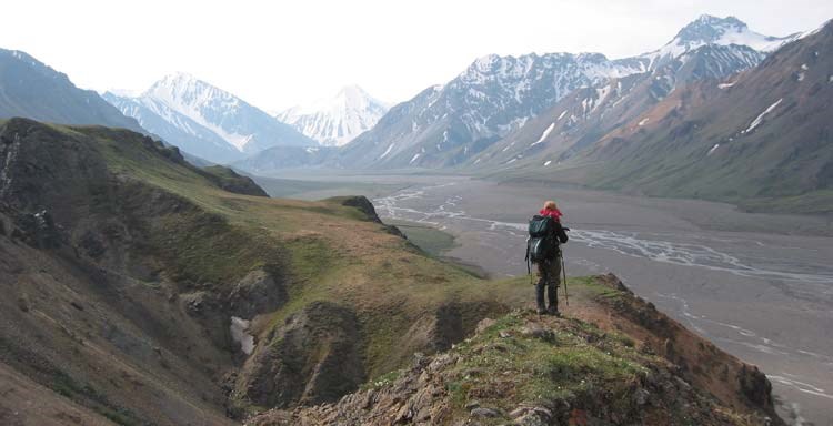 a hiker stands on a rocky ridge, overlooking a wide gravel river bed and distant mountains