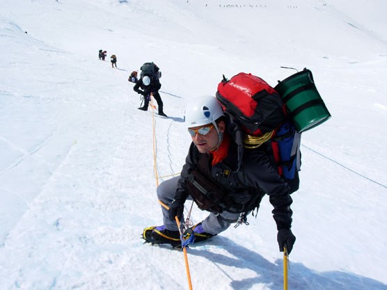 looking down a snowy slope at several mountain climbers, laden with equipment