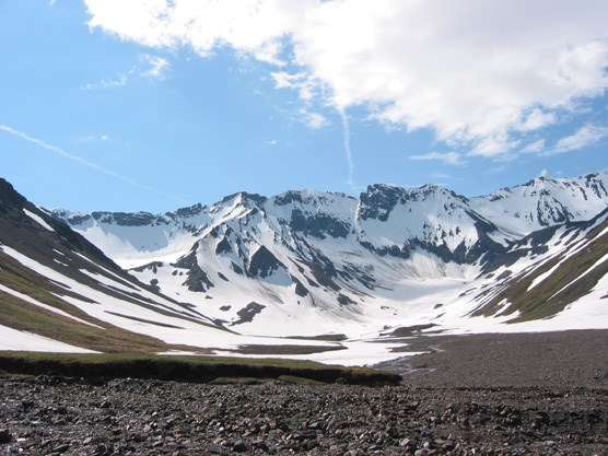 a rocky plain leading up to snow-covered mountains, under a blue sky with one large cloud