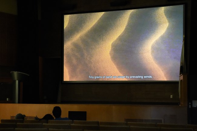 A silhouette of a couple watching the park film in the Furnace Creek Visitor Center Auditorium. The screen shows closed captioning and sand.