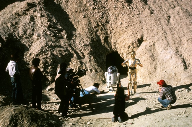 Two robots are surrounded by a few people filming them in a desert landscape.
