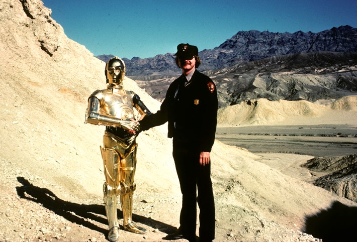A gold robot shakes hands with a park ranger in the desert.