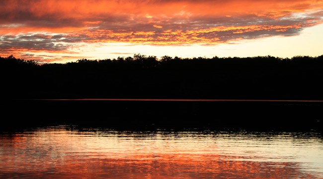 A pink and orange sky over a lake with silhouettes of trees.