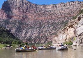 Several rafts float on the river and a canyon wall looms behind them.