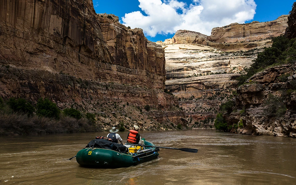Rafting in Lower Yampa Canyon 2 people in a green rubber raft float on a calm river as cliffs of yellowish tan rock tower above them