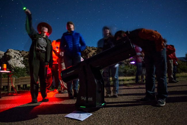 Visitors look into a telescope while a ranger points out deep sky objects with a laser amid a backdrop of red lights.