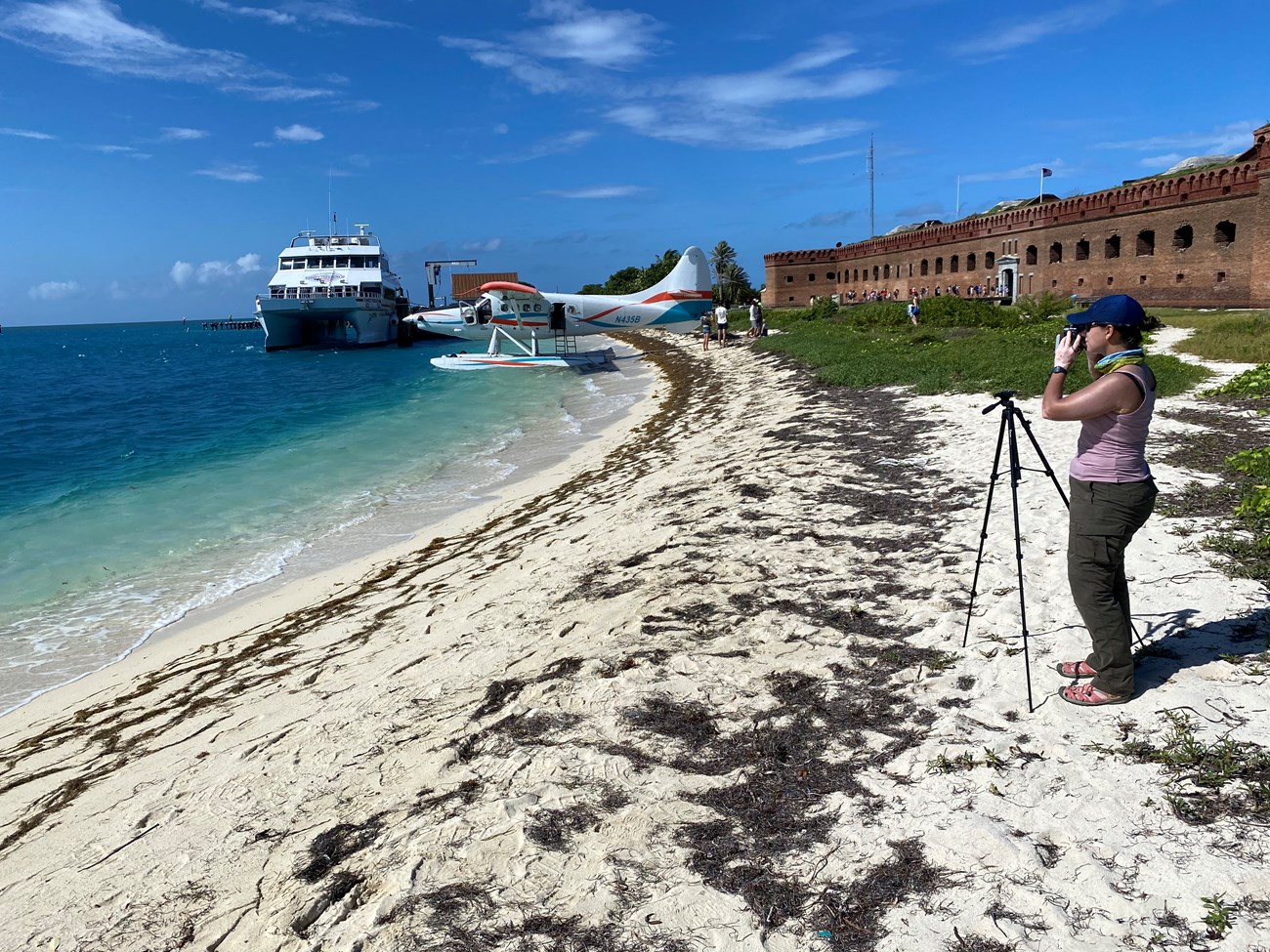 A woman photographing a seaplane and a boat docked by a beach and a large brick structure