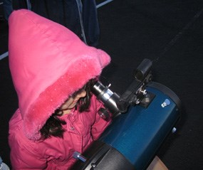 A young girl with a fuzzy pink coat peers into a telescope.