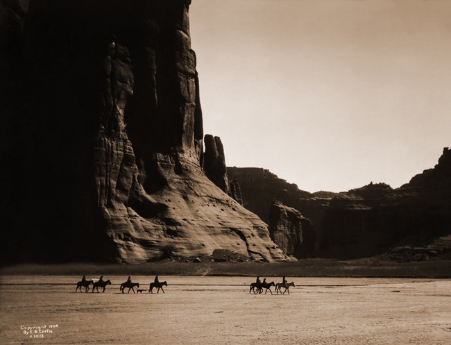 Seven people riding on horseback are dwarfed by the sandstone landscape around them.  A cliff juts up behind four of the riders. (Archival photo)