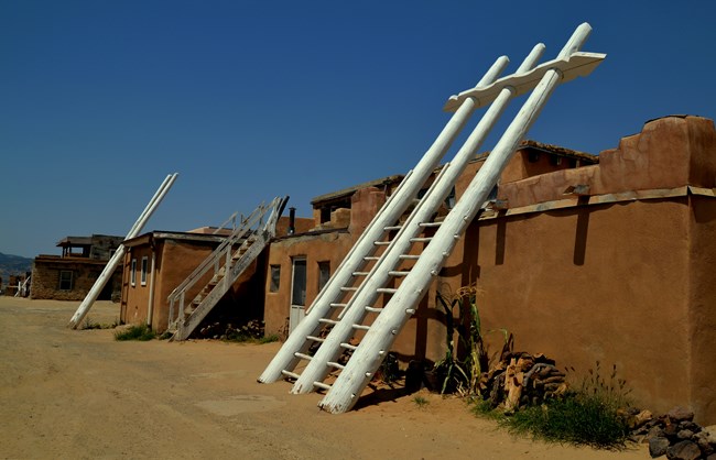 A white, wooden ladder leans against a wall of a single-leveled pueblo. Rungs stretch between the three tree trunks that make its frame. Rungs start at the ground and end at the rooftop, while the ladder's frame stretches far above the roof.