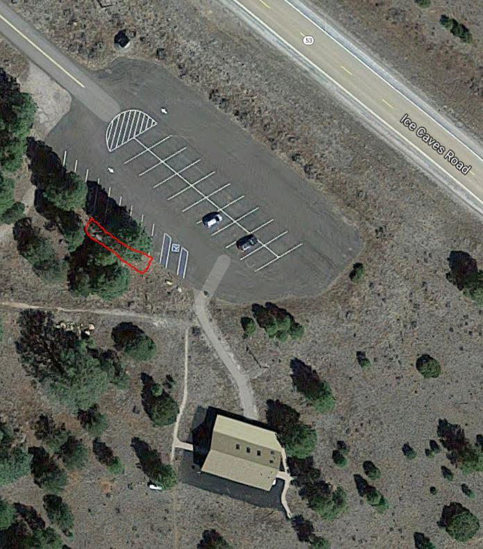 A satellite view of a small building next to a parking lot and road, surrounded by scattered trees.