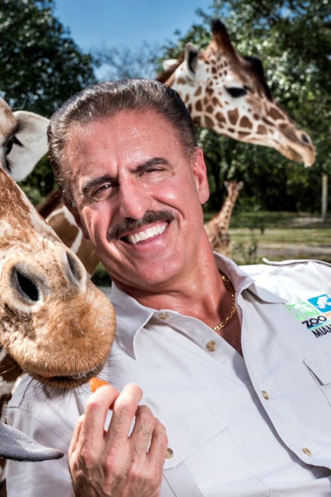 A giraffe leans its snout on the shoulder of a smiling Ron Magill while another giraffe grazes in the background