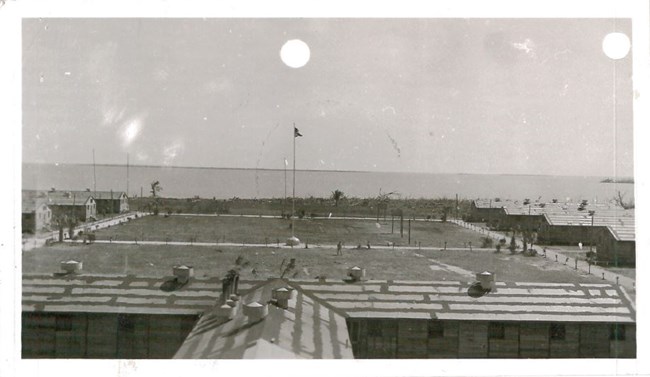 A black and white image of several small barn-like buildings positioned along the perimeter of a rectangular field. There is a flagpole in the center of field and the ocean is visible in the background.