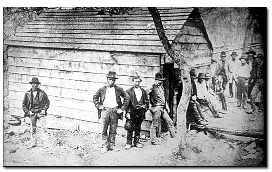 Black and white photo from the late 1870s of a small wooden shack with thirteen men of various races standing next to it. The picture is taken from the side with part of one end showing. No door or window is visible; the roof is gabled with no shingles. There is a small tree growing right next to the end of the structure and the hillside is bare.