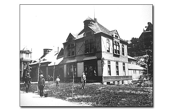 Black and white photo of a two-story structure of eclectic design.  The [south] end nearest the photographer has a hipped roof with two gabled dormers on the front and one of the end. The north half of the building has a gabled roof with an inset porch. There are four men in dress clothes spaced out in front of the building and on the sidewalk. There are small sapling trees next to the sidewalk.
