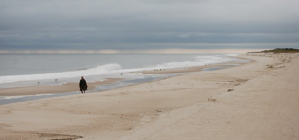 Lone hiker strolls along beach, as rain falls from thick dark clouds on the horizon.